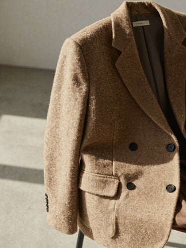 A close-up of a warm oatmeal wool blazer draped over a chair in a sunlit, minimalist concrete studio, soft shadows.