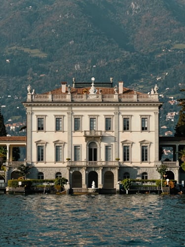 elegant editorial photography, soft natural lakeside light, deep teal and warm white tones, architectural symmetry, medium format. A majestic historic villa on the shores of Lake Maggiore, beautiful stone facade, pristine gardens, calm water in the background.