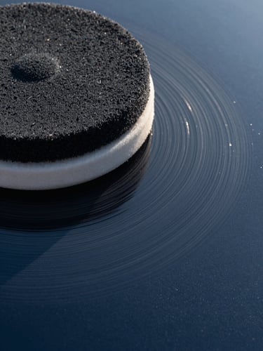 Extreme close-up of a rotary buff pad pressing against freshly polished dark navy car paint, cool studio lighting catching the circular polish trail and surface depth, framed from above-left with the pad filling the right two-thirds of the frame
