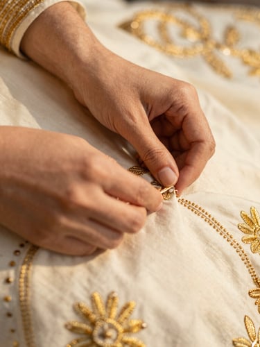 Close-up of hands detailing golden tilla embroidery on ivory fabric, rich textile textures, warm editorial fashion photography, shallow depth of field