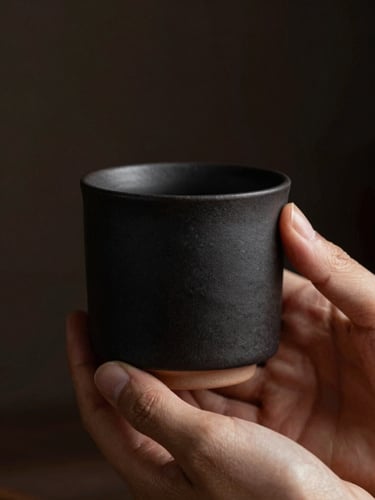 Moody product photography with low-key side lighting. A close-up of a matte-black ceramic cup held by hands, highlighting the raw unglazed texture.