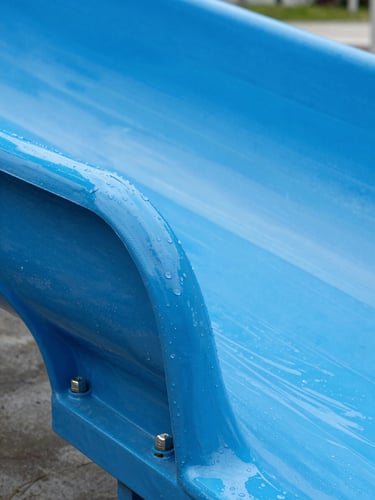 Macro close-up of a heavy-duty commercial water slide joint, showing thick blue fiberglass, polished stainless steel bolts, and clean water droplets, bright daylight