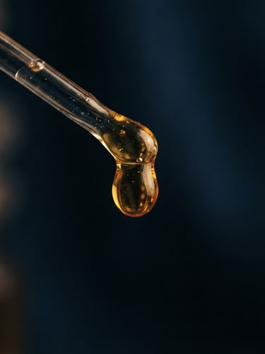 Warm low-key macro shot of a golden Attar droplet suspended from a glass applicator, rich amber liquid, dark navy background, 35mm