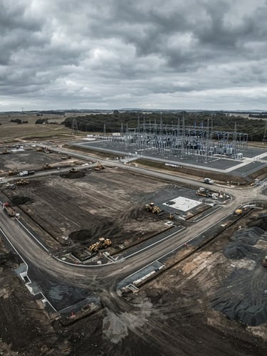 Aerial drone photography of graded land parcel with high-voltage substation in background, overcast dramatic daylight, cool desaturated tones, wide-angle lens