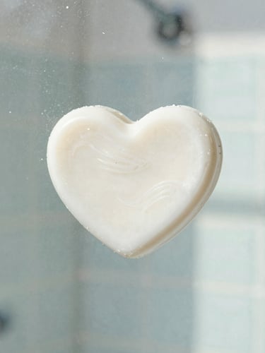 Close-up editorial photography of a hand-drawn soap heart on a sparkling clean glass shower door, warm natural light reflecting off the surface, sharp focus, 35mm lens.