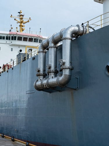 Close-up panoramic of a large tanker vessel's hull and pipeline manifold at an industrial port, steel infrastructure, cool blue-grey industrial light, no people, extreme horizontal framing