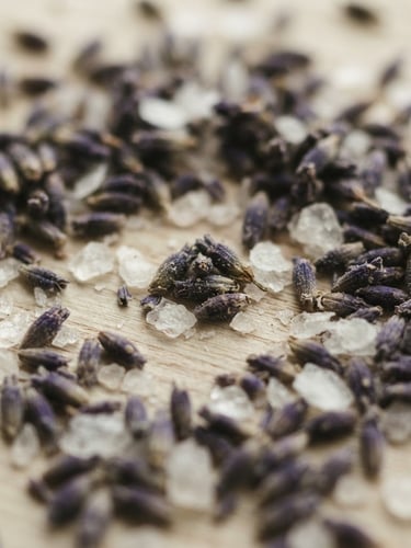 Macro close-up of dried lavender buds and magnesium flakes on a light ash wood surface, soft diffused daylight, shallow depth of field, muted sage and cream tones.