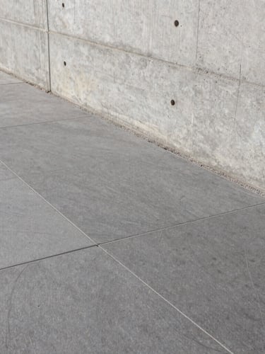 Close-up of premium stone flooring installation next to a raw concrete wall, clean architectural lines, sharp daylight.