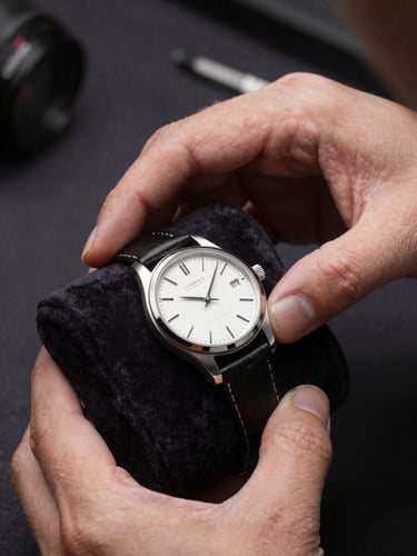 Close-up of hands in a clinical, non-posed manner examining a fine wristwatch on a dark velvet pad under directional studio light, no faces visible, detail of the watch dial sharp and centered