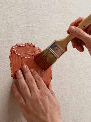 Close-up of hands with paint-stained fingers holding a professional brush, applying thick terracotta-colored paint onto a heavily textured linen canvas, soft natural light