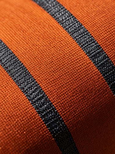 Macro close-up of orange and charcoal technical fire-retardant weave, showing heavy-duty aramid stitching, rugged tactile texture, dramatic side lighting casting deep shadows.