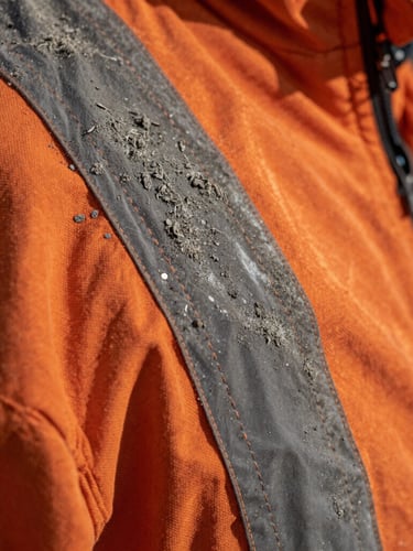 Macro portrait of rugged orange fire-retardant textile, close-up on heavy double-stitching and high-vis reflective strip, dusted with fine grey ash, dramatic side lighting.