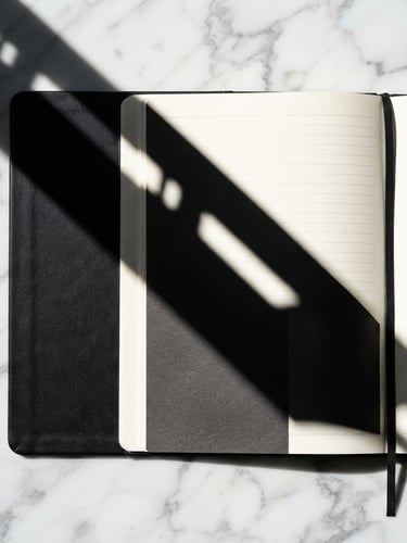 Macro close-up of a sleek physical black notebook resting on a white marble desk, sharp architectural shadow cutting diagonally across the page, 50mm.
