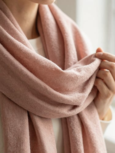 Close-up of hands gently holding a folded dusty rose scarf, showing the fine weave and soft fabric texture, warm natural window light