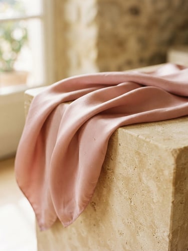 Close-up of elegant dusty pink silk scarf draping softly over a warm beige stone pedestal, diffused natural window light, editorial photography, 35mm