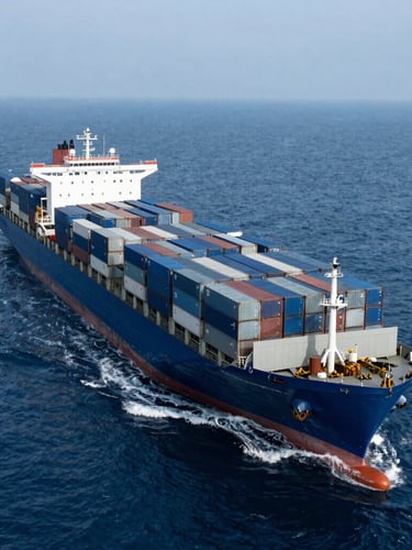 Wide-angle overhead shot of a massive container ship loaded with blue and gray cargo containers navigating the deep blue open ocean under cool overcast daylight.