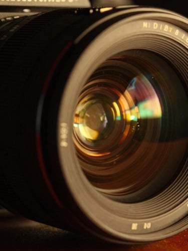 Anamorphic close-up of a high-end film camera lens reflecting golden stage lights, deep shadows, cinematic atmosphere