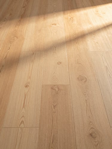 Wide close-up of freshly installed wide-plank white oak hardwood flooring filling the right frame edge to edge, warm golden-hour side light raking across the grain and seam lines, extreme horizontal framing, no people, no furniture