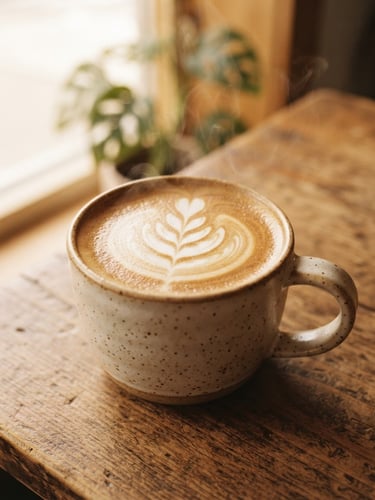 Close-up of a steaming ceramic mug of latte art on a warm wooden table, soft morning window light, rich espresso crema, shallow depth of field, 35mm
