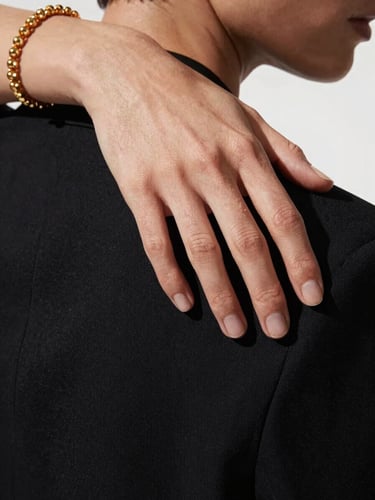 Extreme close-up of a model's hand resting on the shoulder of a structured black blazer, editorial lighting with a defined shadow cutting diagonally across the fabric texture, warm gold jewellery detail visible at the wrist, stark white background