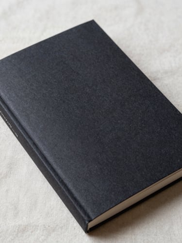 Close-up of a single novel held open at an angle on a cream linen surface, soft north-facing daylight from above-left, the cover faces the viewer — dark minimal Indonesian design, paper texture visible at the spine, no flash