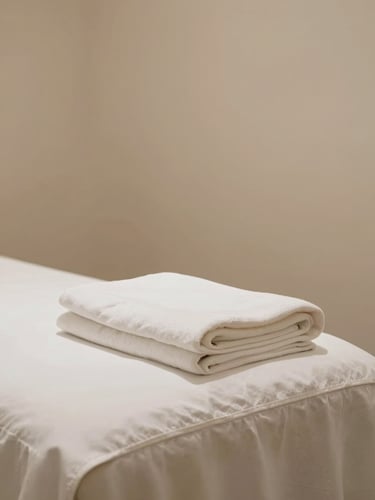 Serene empty treatment room with crisp white folded linen on a massage table, soft warm ambient lighting, minimalist clinical-zen atmosphere.