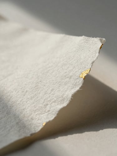 Macro close-up of deckle-edged cotton paper with gold foil details, soft-focus studio morning light, long shadows.