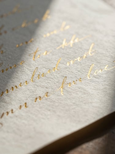 Macro close-up of deckle-edged cotton paper with delicate gold foil calligraphy, soft natural morning light, long gentle shadows