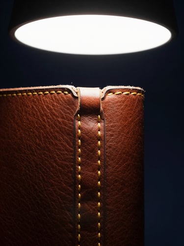 Tight close-up of a hand-stitched leather binding, gold thread visible at the seam, controlled studio strobe light from above, dark navy background, symmetrical framing