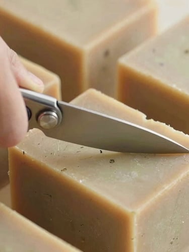Close-up of a hand cutting a large block of natural herbal soap with a wire cutter, botanical flecks visible in the soap texture, warm daylight
