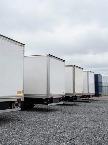 Wide environmental shot from ground level, a row of clean semi-trailers staged in a gravel yard under flat overcast daylight, trailers aligned and ready, no drivers present, steel and aluminum surfaces catching diffuse grey light, industrial fence visible at far edge