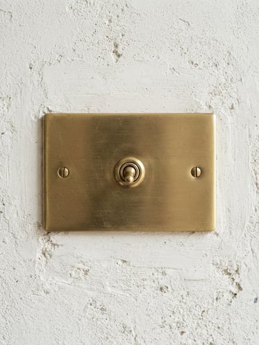 Bright architectural photography of a premium brushed brass light switch mounted on a textured off-white plaster wall. Soft natural daylight casting gentle shadows, sharp macro detail showing the metal grain, 35mm lens, straight-on architectural angle.