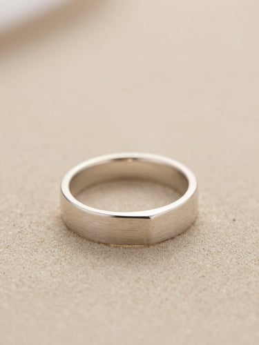 Macro product photography of a hand-finished brushed silver ring with clean geometric edges, soft diffused daylight, warm sand background