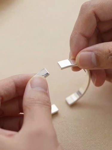 Extreme close-up of a craftsman's hands carefully polishing a solid silver geometric cuff bracelet, soft daylight, warm sand background, macro lens