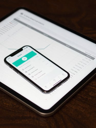 Tight macro shot of an iPad displaying a clean financial spreadsheet and an iPhone showing an ad manager dashboard on a dark walnut desk, sharp side lighting, deep shadows