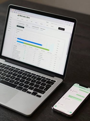 Close-up overhead flat-lay of a laptop screen showing a Mercado Libre product listing dashboard with analytics chart, on a dark desk with a phone showing WhatsApp conversation beside it, clinical studio lighting