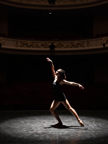 Wide environmental shot of Victoria Holanda in mid-movement on a dark empty theater stage, high-contrast stage lighting, dramatic shadows, muted monochrome with warm undertones, 35mm.