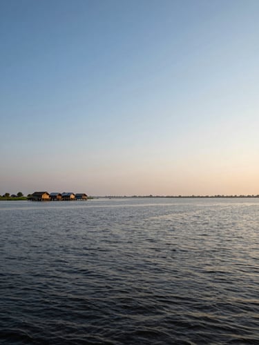 Wide environmental shot of the haor wetlands in monsoon fullness — water reaching to every horizon, a small cluster of homesteads raised on earthen mounds visible in the far left third, late afternoon light casting long gold lines across the water surface, shot from a low angle at water level emphasising the vast open sky above