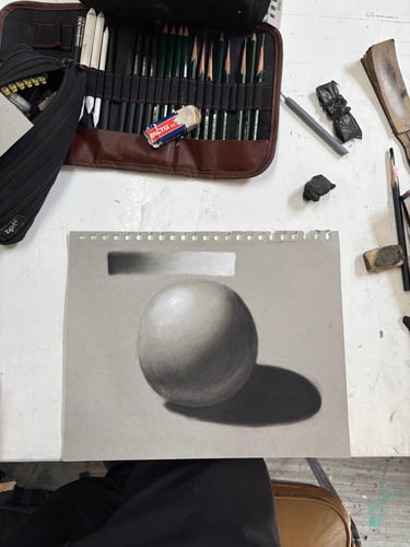 Shaded pencil drawing of a sphere on gray paper with art supplies and drawing pencils.