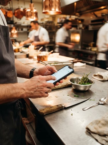 Close-up of hands using a mobile device on a stainless steel prep table in an active kitchen, warm ambient lighting, sharp focus, 35mm
