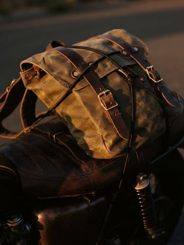 Close-up of a rugged waxed canvas backpack strapped to the leather seat of a custom motorcycle, warm sunset light hitting the metallic buckles, rich textures, moody film photography