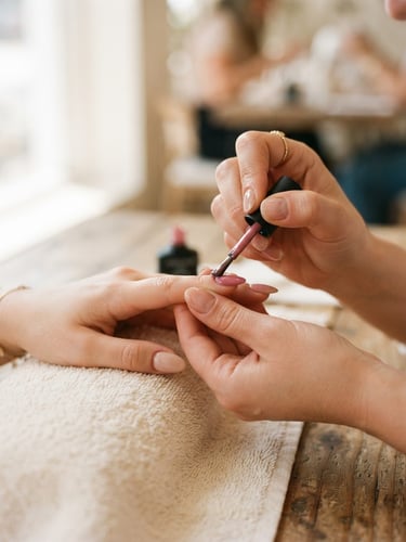 Close-up of a manicurist's hands meticulously applying gel polish to a client's nail, with soft natural window light illuminating the scene, shallow depth of field, rich cream and pink tones.