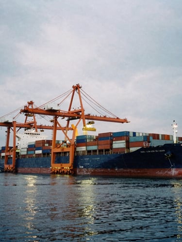 Industrial film photography of a massive container ship docked at a deep-water port during overcast twilight, giant orange gantry cranes loading cargo, reflections on dark water, wide-angle lens.