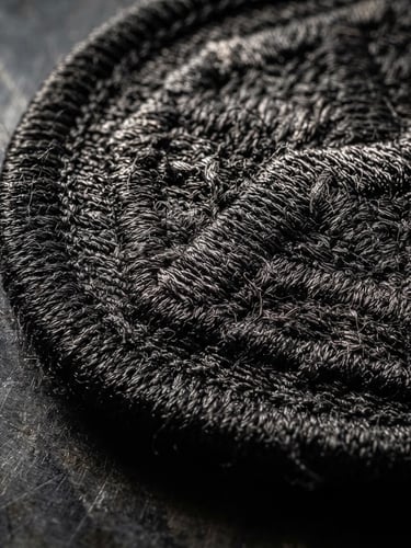 Tactile macro photography, hard directional side-lighting, extreme close-up of heavy-duty embroidered patch with a thick merrowed border on a dark steel background, showing dense thread weave and sharp physical texture.