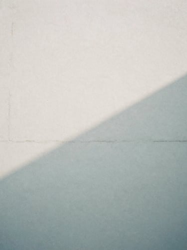 Abstract architectural shadow falling across a pale concrete wall, soft overcast daylight, desaturated cool tones, high grain, 35mm lens, quiet atmosphere