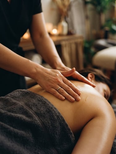 Close-up of a female therapist's hands gently applying oil to a client's draped shoulder, soft golden ambient light, shallow depth of field, pristine treatment room in background, 35mm
