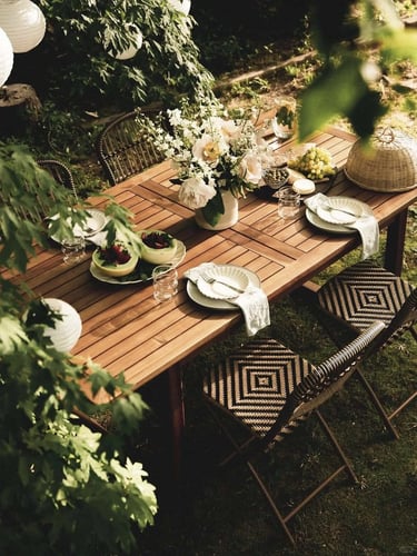 Outdoor garden dining area with wooden table, chairs, hanging lights and lush greenery creating a pe