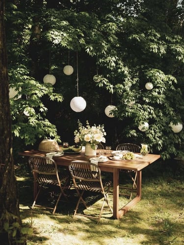 Outdoor garden dining area with wooden table, chairs, hanging lights and lush greenery creating a pe