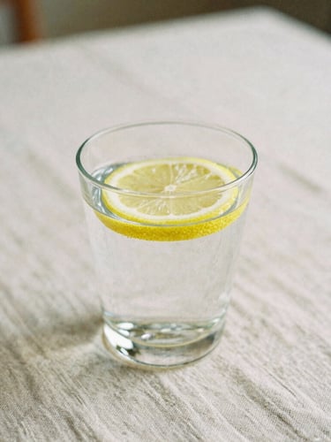 Medium shot of a glass of clean water with a lemon slice on an organic linen tablecloth, soft morning light, shallow depth of field