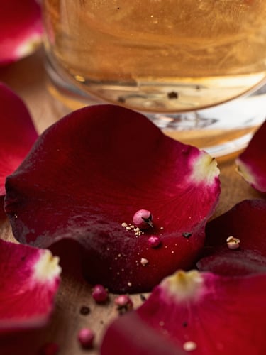 Close-up macro of velvety dark red rose petals and crushed pink peppercorns, sharp glass reflections in the background, warm golden hour glow, deep contrast